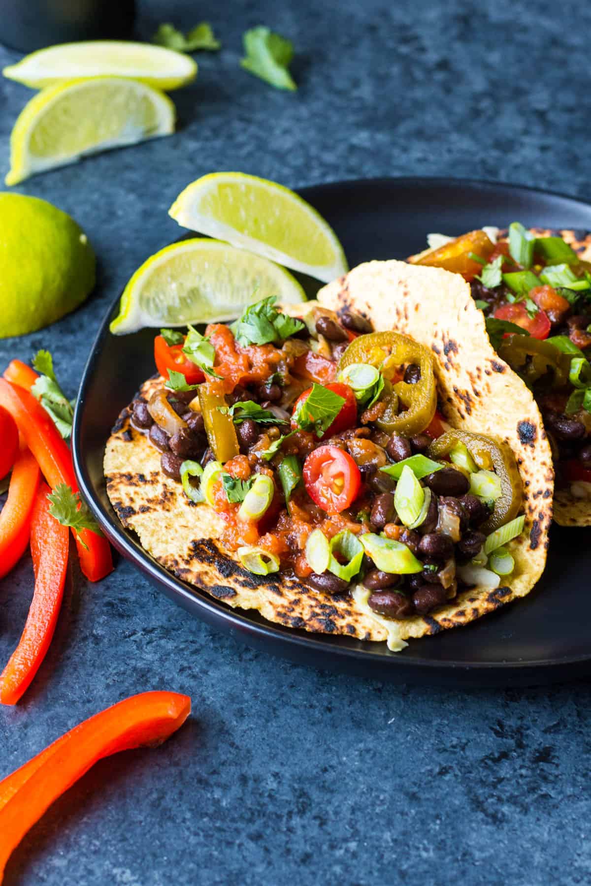 black bean tacos with vegan creamy slaw on a black plate with salsa, tomatoes, green onion, and lime