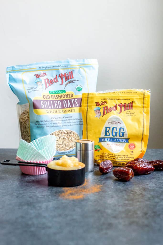 A bag of Bob's Red Mill Rolled Oats, a bag of Bob's Red Mill Egg Replacer, applesauce, muffin liners, dates, and cinnamon